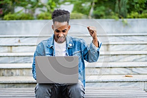 Happy successful african-american man with arms raised in air. Young casual student with laptop celebrating victory outdoors,