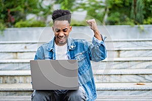 Happy successful african-american man with arms raised in air. Young casual student with laptop celebrating victory outdoors,