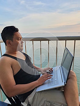 Happy successful Asian man using laptop while sitting on his balcony with calm blue ocean behind, remote working digital nomad