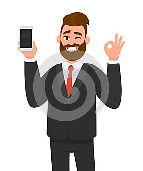 Happy successful businessman showing smart phone, mobile, cell phone in hand and gesturing okay or OK sign while winking. Good.