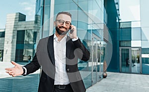 Happy successful businessman talking on smartphone, standing outdoors against modern office center, free space