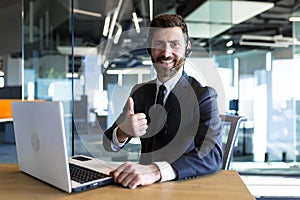 Happy and successful call center employee looks at the camera and smiles, businessman keeps his finger approvingly up, working at