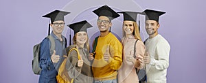 Happy successful university students in black caps giving thumbs up on purple background