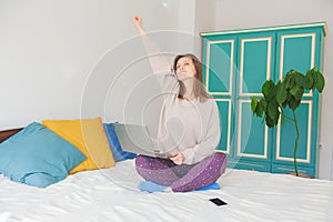 Happy successful woman sitting on the bed and working with computer and smartphone at home with hand up