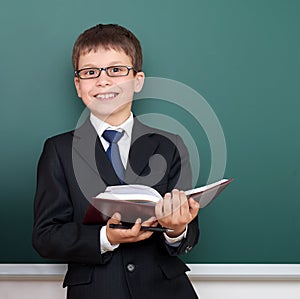 Happy successfull school boy with book portrait, dressed in classic black suit, on green chalkboard background, education and