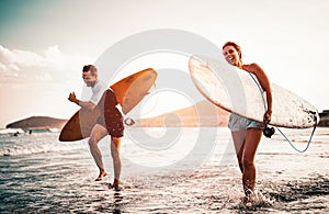 Happy surfer couple running with surfboards along the sea shore - Sporty people having fun going to surf together at sunset