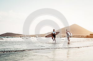Happy surfer couple running with surfboards along the sea shore - Sporty people having fun going to surf together at sunset