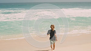 Happy surfer with curly long hair running on sand beach to idyllic ocean coast with waves. Male hippie traveler on