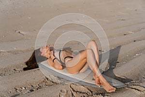 Happy surfer girl in swimsuit lying on surfboard at sandy beach of ocean.