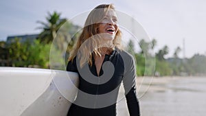 Happy surfer girl in swimsuit walking along beach carrying surfboard, smiling, talking, flirting with boyfriend