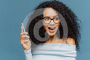 Happy surpirsed African American woman holds on cell phone, being interacted by telephone call, focused aside happily, opens mouth