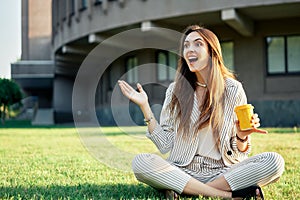 Happy and surprised beautiful woman look away sitting on green grass with coffee