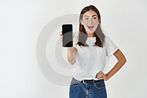 Happy and surprised brunette woman showing blank smartphone screen at camera, standing over white background