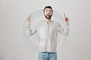 Happy surprised european man pointing and looking up, staring with opened mouth and standing against gray background