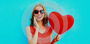 Happy surprised woman laughing with bunch of red heart shaped balloons wearing a sunglasses on a blue background