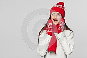 Happy surprised woman looking sideways in excitement. Excited christmas girl wearing knitted warm hat and scarf, isolated on gray