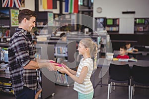 Happy teacher giving book to schoolgirl in library