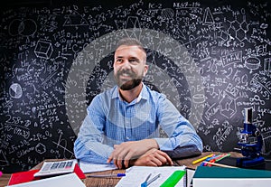 Happy teacher sitting at desk, school supplies, big blackboard