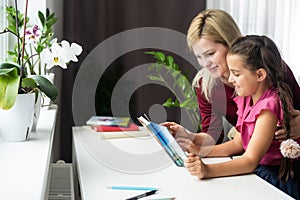 Happy teacher and student girl sitting at working desk in classroom. Cheerful young mother and child sitting at table