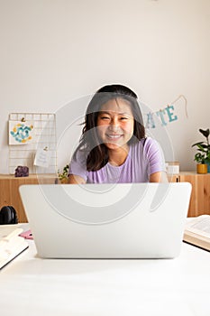 Happy teen asian female high school student studying at home, doing homework using laptop looking at camera. Vertical.