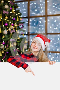 Happy teen girl in christmas hat peeking above white banner and pointing down. Space for text