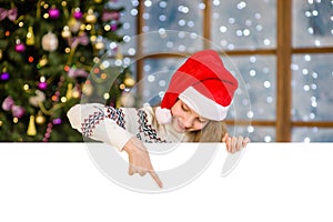 Happy teen girl in christmas hat peeking and pointing down at empty board. Space for text