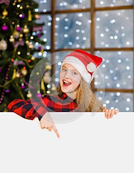 Happy teen girl in christmas hat peeking and pointing down at empty board. Space for text