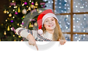 Happy teen girl in christmas hat peeking and pointing down at empty board. Space for text