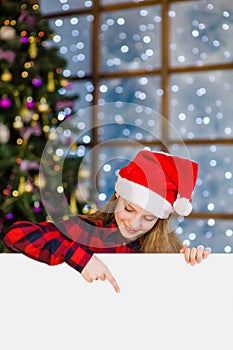 Happy teen girl in christmas hat peeking and pointing down at empty white board. Space for text