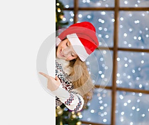 Happy teen girl in christmas hat peeking and pointing at empty white banner