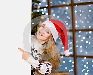 Happy teen girl in red christmas hat peeking above white banner and pointing away