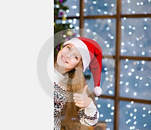 Happy teen girl in red santa hat behind white board and showing thumbs up. Space for text