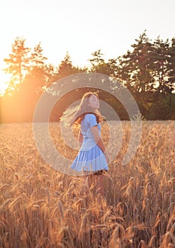 Happy teen girl is smiling at wheat field, touching ears of wheat with her hand. Beautiful teenager enjoying nature in warm