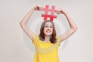 Happy teen girl with toothy smile in yellow t-shirt holding red hashtag sign board on head, tagging photo