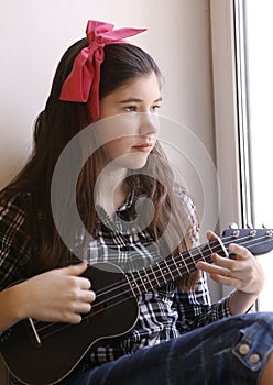 Happy teen girl with ukulele guitar in checked shirt and jeans playing look at the window
