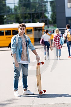 happy teen schoolboy with skateboard standing in front of school bus and group of classmates walking blurred