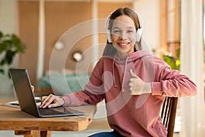 Happy teen schoolgirl in headset gesturing thumb up, sitting at desk near laptop, having online lesson from home