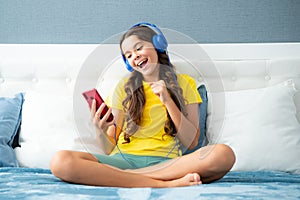 Happy teen teenage girl in headphones relax on bed at home listen to music using laptop. Child in earphones browse