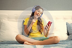 Happy teen teenage girl in headphones relax on bed at home listen to music using laptop. Child in earphones browse