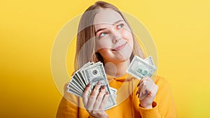 Happy teenage blonde girl holding in hands money cash dollars dreaming thoughtfully isolated on color yellow background