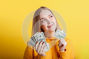 Happy teenage blonde girl holding in hands money cash dollars dreaming thoughtfully isolated on color yellow background. Portrait