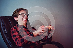 Happy teenage boy playing video games seated in his armchair. Excited adolescent wearing eyeglasses, holding joystick console