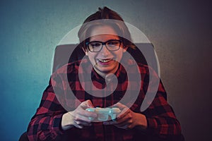 Happy teenage boy playing video games seated in his armchair. Excited adolescent wearing eyeglasses, holding joystick console