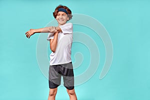 Happy teenage boy in sportswear looking away and smiling while stretching his arms before workout or training, standing