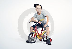 Happy teenage boy in a T-shirt, cap and shorts is sitting on a bicycle, on a light background. File contains a path to isolation