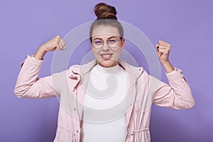 Happy teenage female posing against lilac studio wall clenching fists and celebrating her success, looking smiling directly at