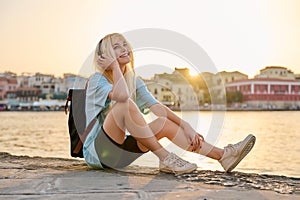 Happy teenage female tourist sitting on sea promenade of an old city in sunset