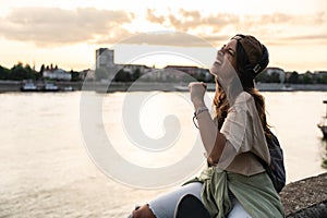 Happy teenage female tourist sitting on sea promenade of an old city in sunset. Young woman world traveler singing her favorite