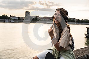Happy teenage female tourist sitting on sea promenade of an old city in sunset. Young woman world traveler singing her favorite
