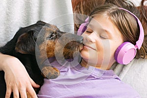 Happy teenage girl in lilac t-shirt in pink headphones listens to music and has fun playing with dog